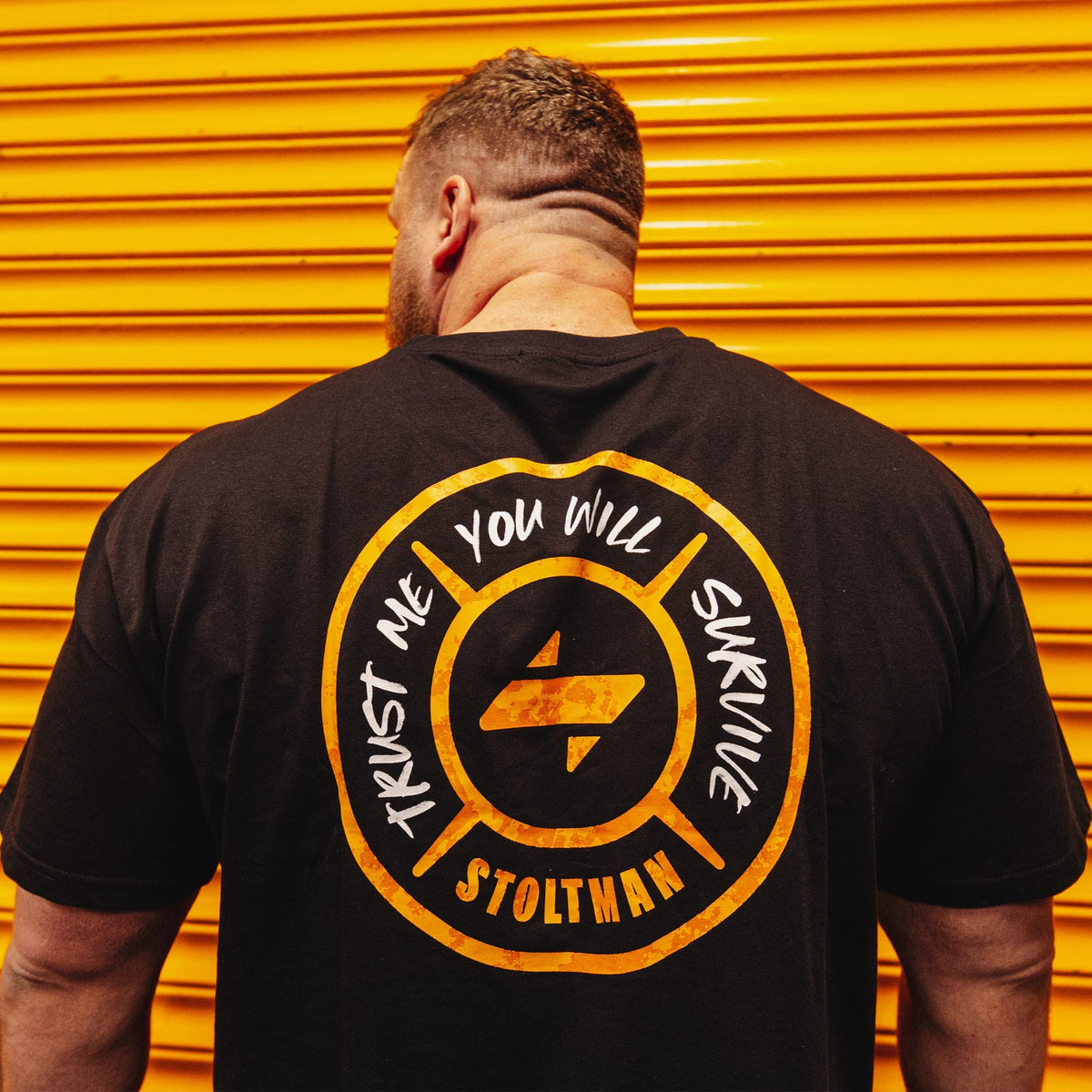 A man viewed from behind wearing a black t-shirt with a large yellow and white circular graphic on the back, standing in front of a bright yellow-orange corrugated metal wall. The graphic features the text 'TRUST ME YOU WILL SURVIVE' and 'STOLTMAN' around a central logo.
