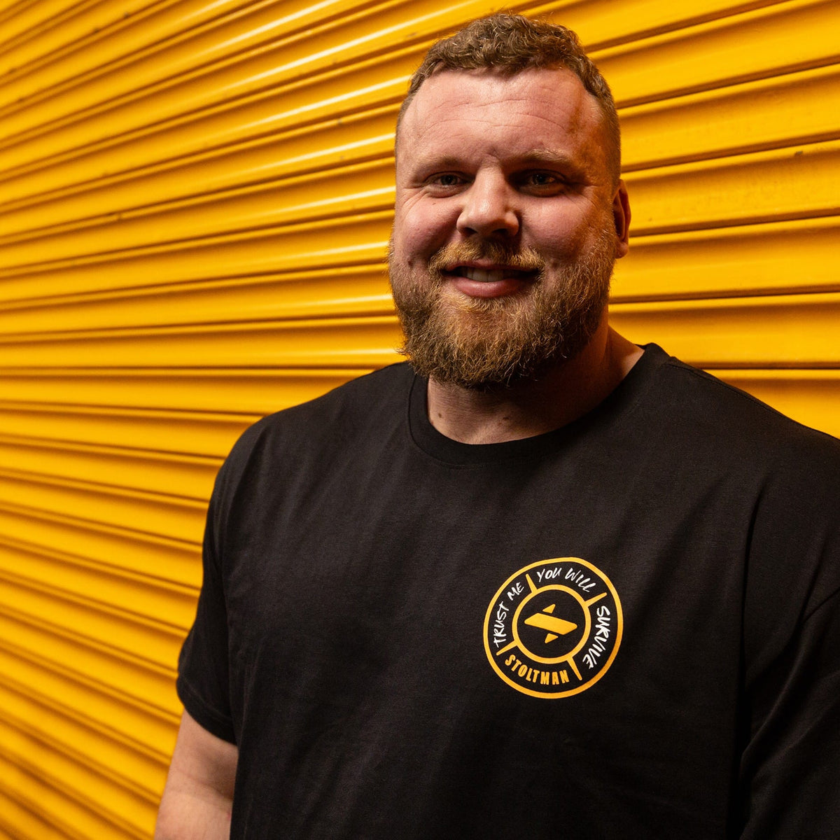 A man with a beard wearing a black t-shirt with a yellow circular logo, standing in front of a bright yellow corrugated metal wall.