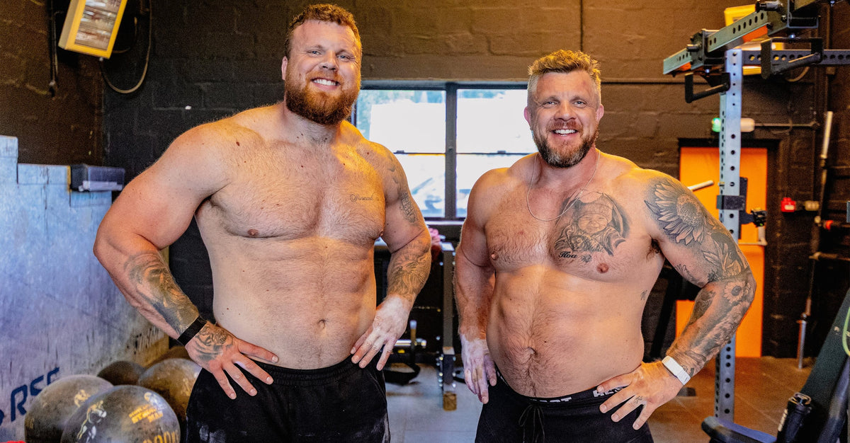 Two shirtless, tattooed men standing side by side in a gym setting, wearing black shorts with chalk dust on them. The background shows gym equipment, weight balls, and a dark brick wall.