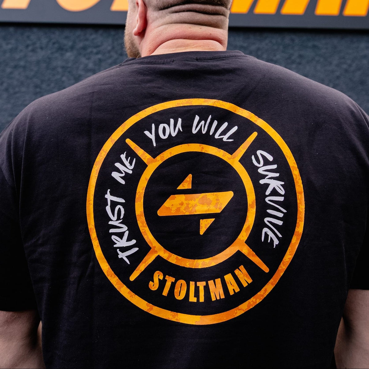 A man viewed from behind wearing a black Stoltman t-shirt with a large yellow and white graphic on the back, standing in front of a dark textured wall with a large yellow Stoltman logo.