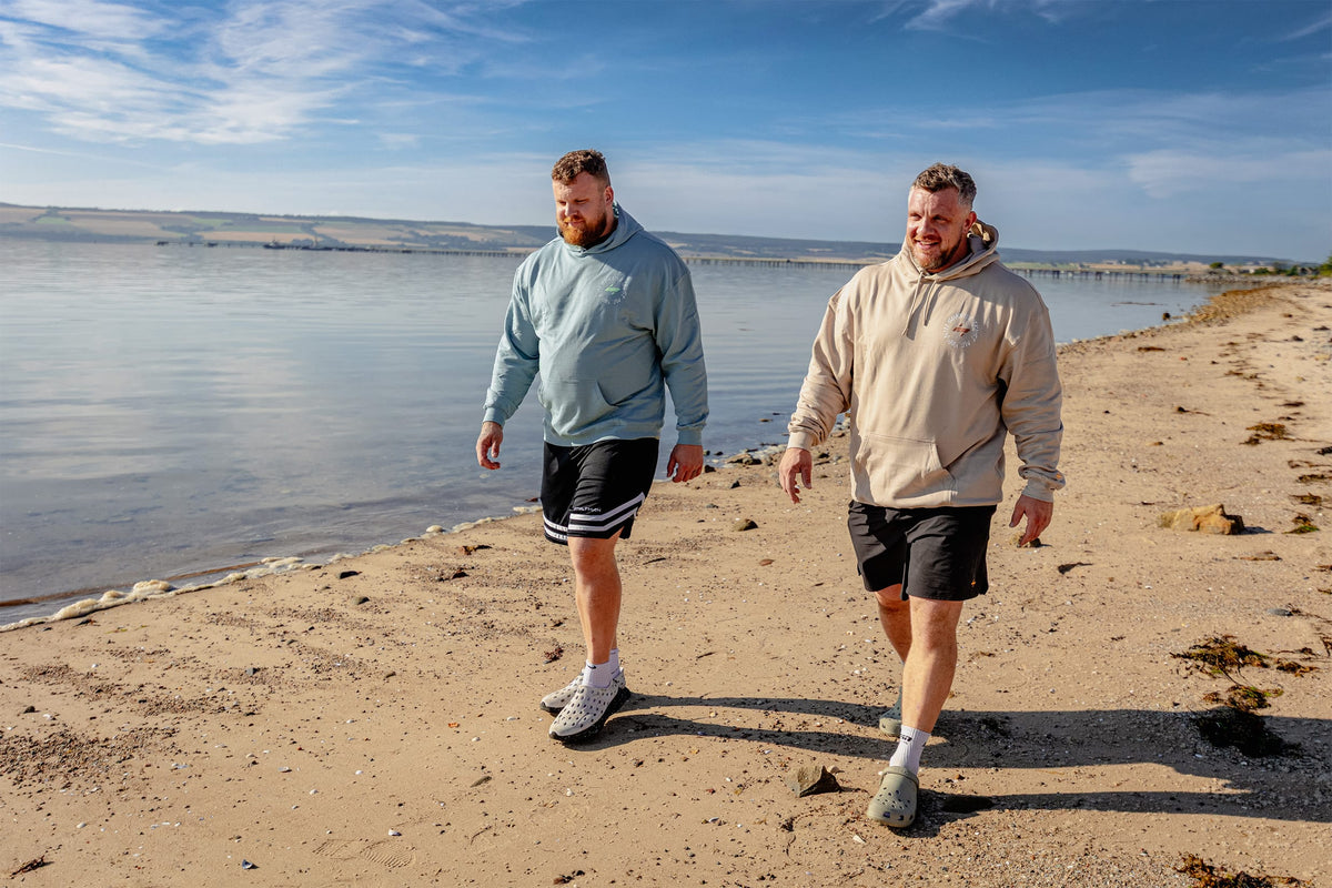 Two men walking on a sandy beach next to a calm lake under a blue sky. The man on the left wears a light blue hoodie and black shorts, while the man on the right wears a beige hoodie and black shorts.
